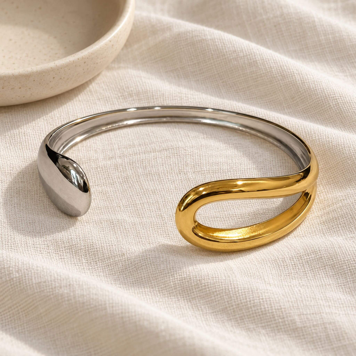 Gold and silver sculptural cuff bracelet on ivory linen beside a ceramic dish, showing its gold-tone open loop and silver-tone end.