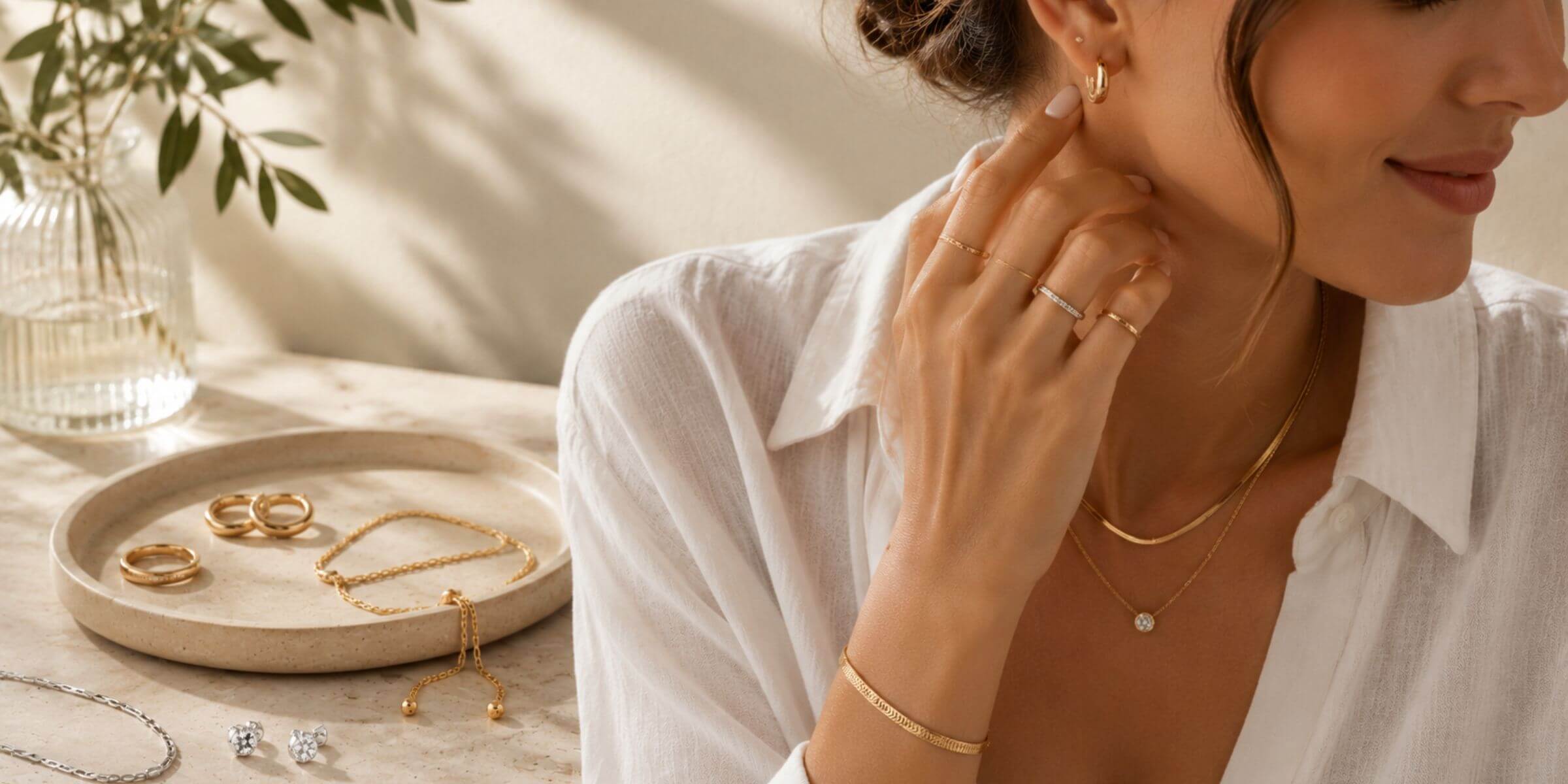 Woman wearing minimalist sterling silver and 18K gold-plated stainless steel jewelry with necklaces, earrings, rings and bracelet in a bright luxury lifestyle setting.