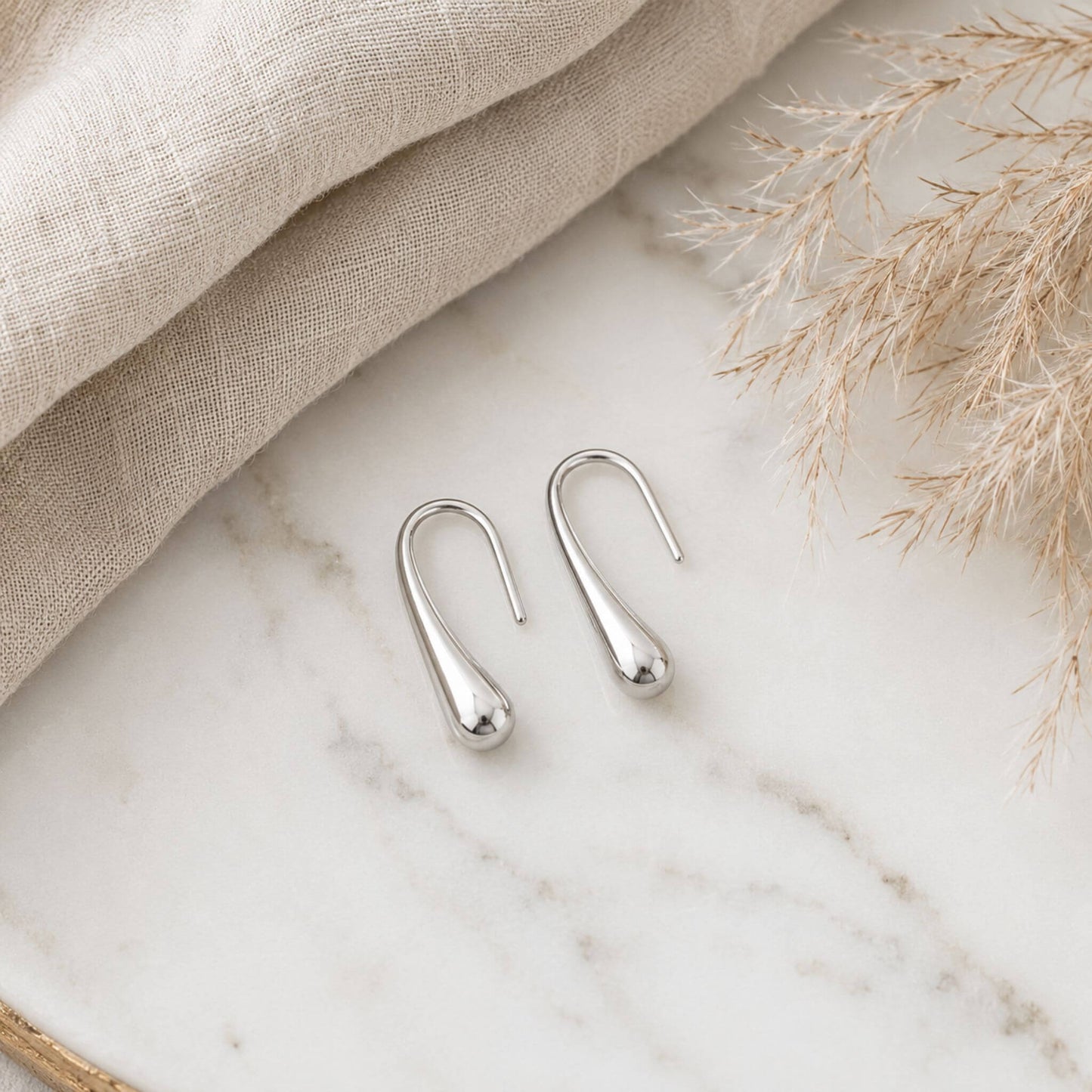 Polished silver stainless steel teardrop hook earrings displayed on a marble surface with neutral linen fabric, lightweight hypoallergenic earrings for everyday wear.